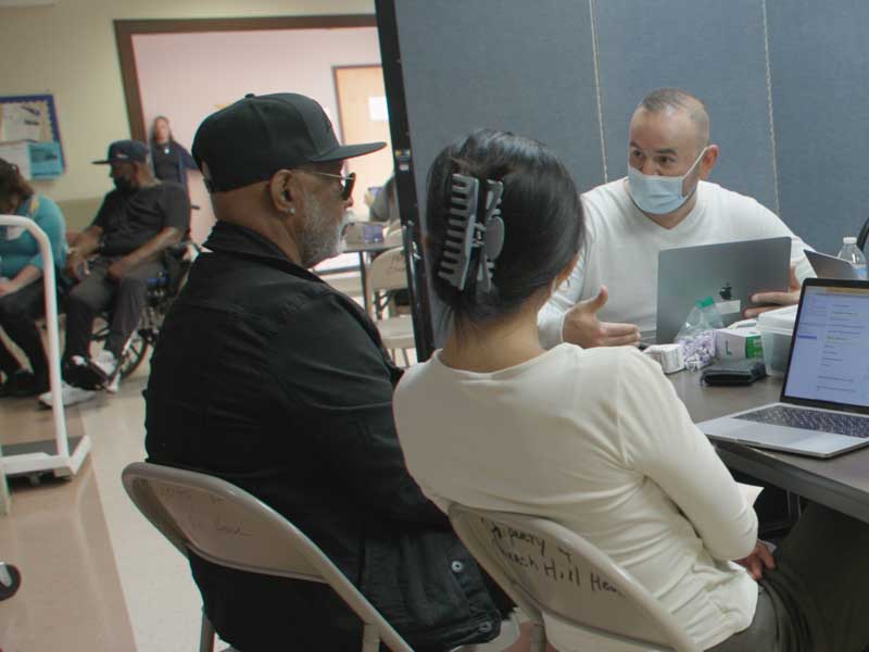 two health care workers with laptops have a discussion with a patient in a health clinic