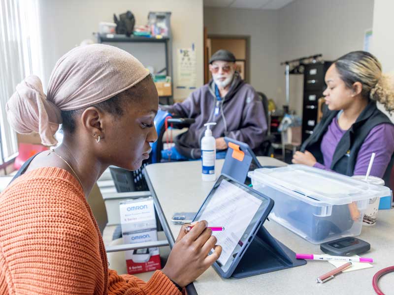 a health care worker in a community clinic enters information into a tablet computer while another worker consults with a patient