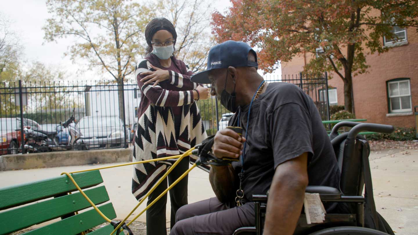 a person in a wheelchair performs arm strengthening exercises with resistance bands attached to an outdoor bench at the behest of a community health worker giving guidance