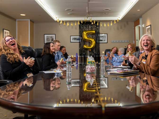 People seated around a conference table laugh and clap during a celebration, with a large gold “5” balloon in the background.