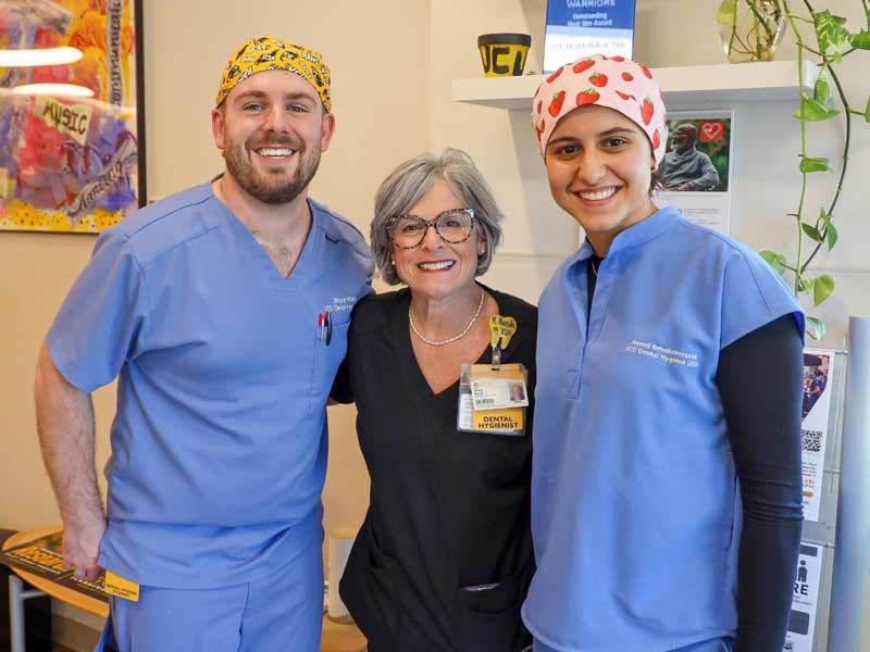 Two students wearing scrubs and patterned surgical caps stand beside a faculty member in scrubs inside a building, all smiling at the camera