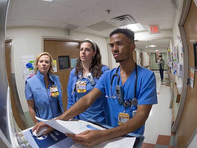 Three nurses in scrubs review a patient chart at a computer workstation in a hospital hallway