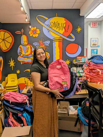 A young woman stands in a room holding a pink backpack, surrounded by tables and stacks of backpacks, with a colorful mural on the wall behind her