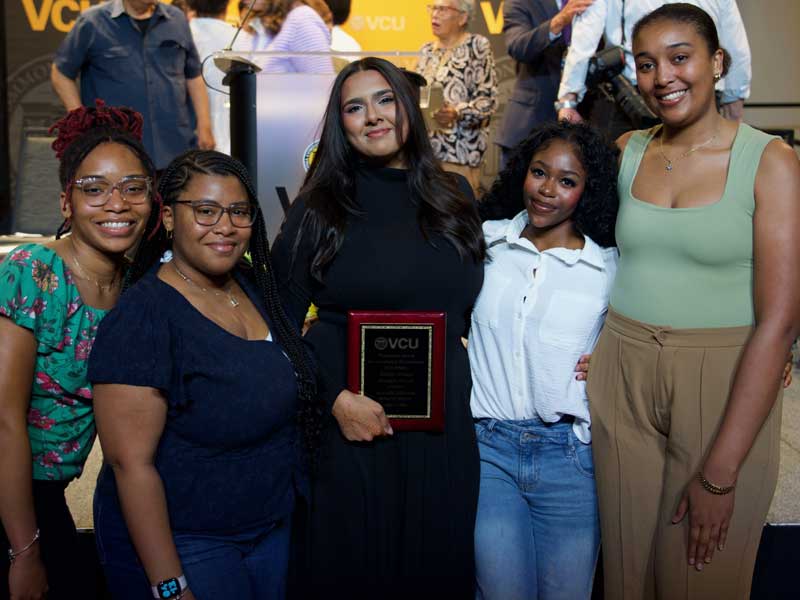 Five students stand together at a V.C.U. event, with one student holding a plaque bearing the V.C.U. logo. A podium and several people are visible in the background.